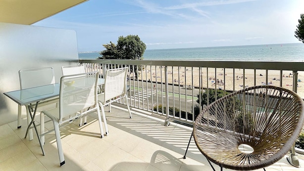A balcony with a glass table and chairs, overlooking a beach with people and a clear sky.