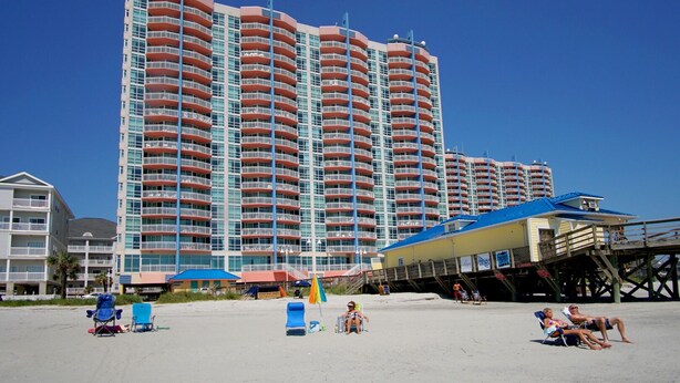 Ein Hochhaus-Hotel mit zahlreichen Balkonen, ein Strandbereich mit Liegestühlen und einer Pier.