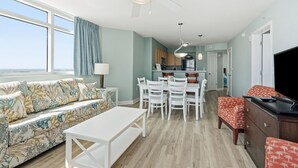 Una sala de estar con un sofá, una mesa de comedor, sillas, un televisor y una cocina con refrigerador y armarios.