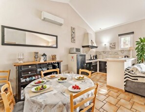 Un comedor con una mesa dispuesta para cuatro, una cocina al fondo y una sala de estar con un sofá y una planta.