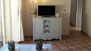 Un saló amb un televisió de pantalla plana sobre un moble blanc, una taula de menjador amb dos objectes decoratius i un passadís que condueix a una altra habitació.