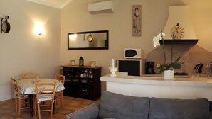 Un saló amb un sofà gris, una taula de cafè blanca, una zona de menjador amb una taula i cadires, un moble negre i un rellotge penjat a la paret.