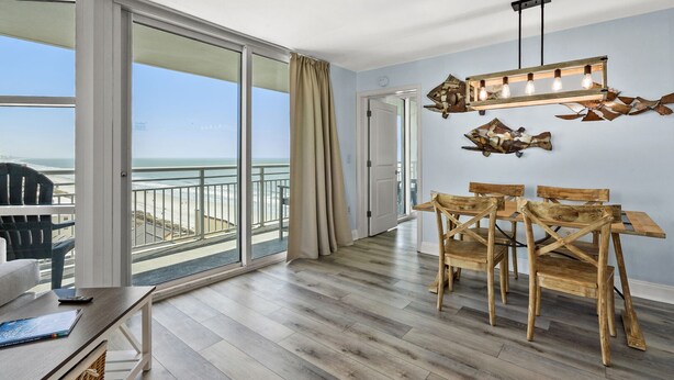 Een ruime kamer met een grote schuifglazen deur die uitkomt op een balkon met uitzicht op de oceaan. Er staat een houten eettafel met stoelen, en erboven een hanglamp.