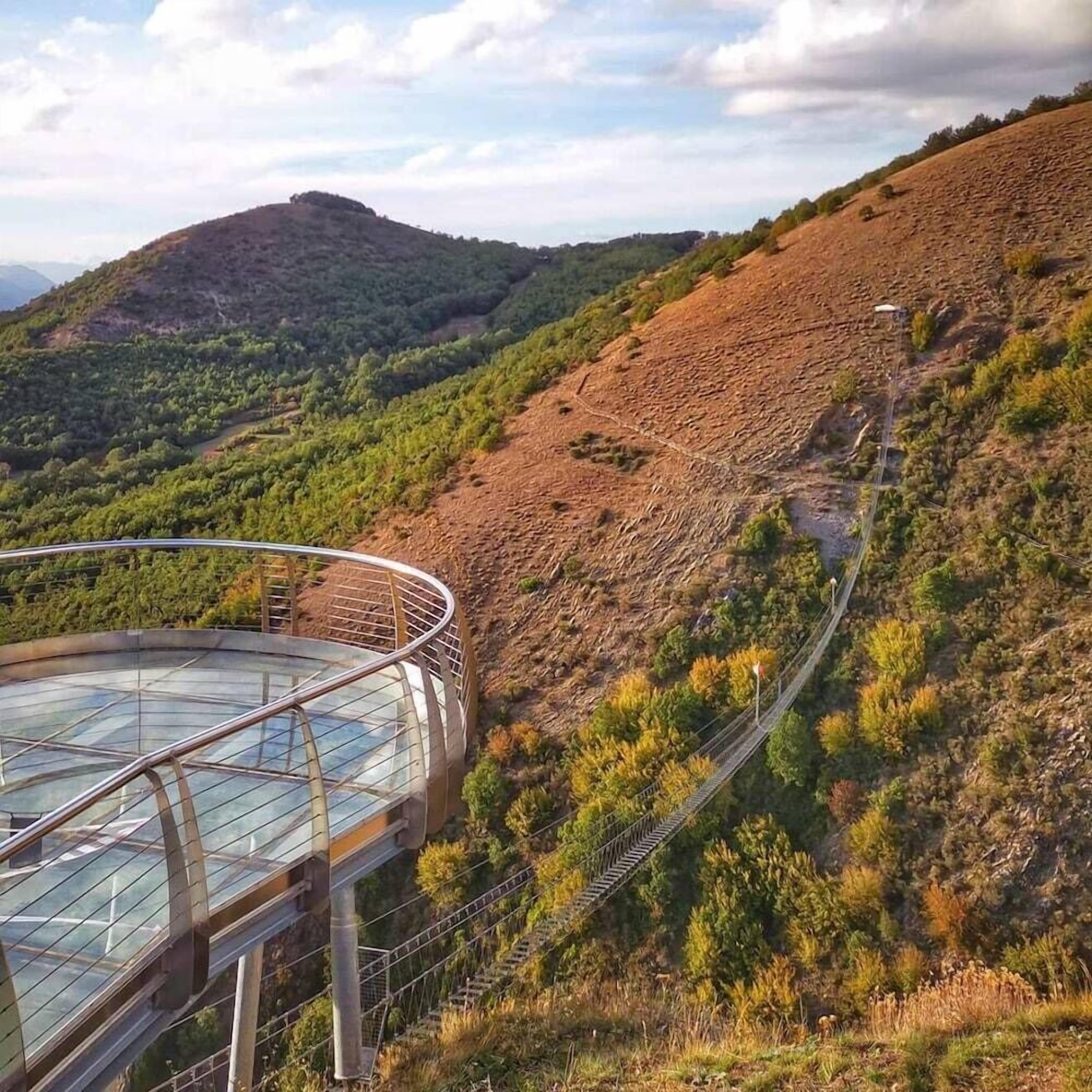 A glass-enclosed observation deck with a metal railing, overlooking a mountainous landscape with a winding road and dense vegetation.
