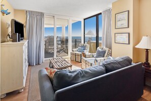 Ein geräumiger Wohnbereich mit einer Couch, gestreiften Sesseln, einem kleinen Tisch und einem Fernseher auf einem Schrank. Durch die Glastüren ist ein Balkon mit Meerblick zu sehen.