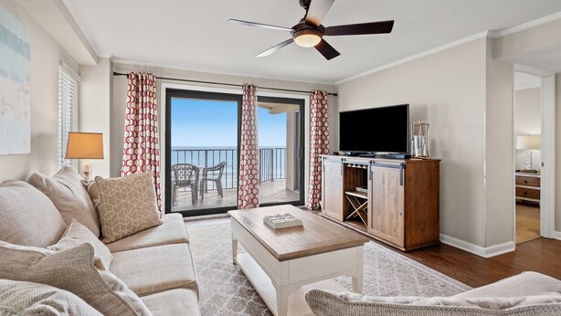 Sala de estar com sofá, mesa de centro e rack para TV. Há uma varanda com cortinas e vista para o mar.