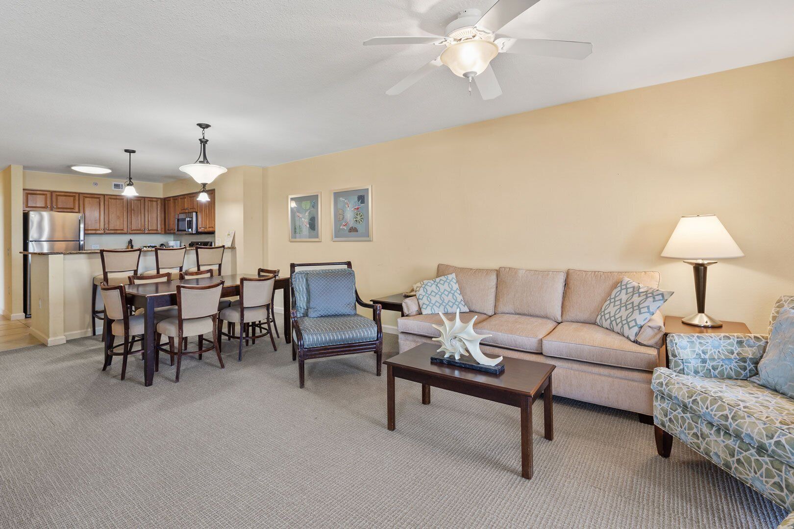 A living room with a sofa, armchairs, a coffee table, and a dining area with chairs and a table. There is a ceiling fan and a lamp on a side table.