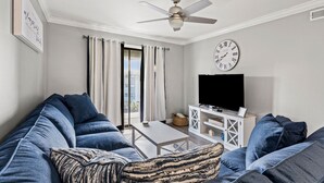 A living room with a large blue sofa, a small table, a TV on a white stand, and a ceiling fan. - Family & fishermen-friendly condos at quiet resort with pool and pier (North Myrtle Beach)