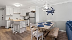 A compact kitchen with white cabinets, a built-in oven, and a refrigerator. There is a wooden dining table with blue placemats and white chairs. - Family & fishermen-friendly condos at quiet resort with pool and pier (North Myrtle Beach)
