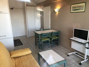 A compact room with a kitchenette, a dining table with chairs, a small sofa, a TV on a stand, and a framed picture on the wall.