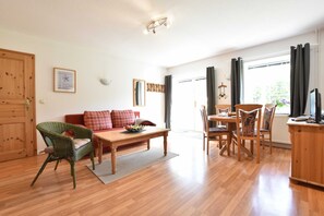 A spacious living room with a wooden floor, a sofa, a dining table with chairs, and a kitchen area with a counter and cabinets.