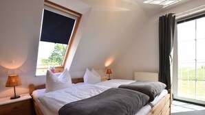 Une chambre avec une grande fenêtre, un lit avec une literie blanche et grise, une table de chevet avec une lampe, et une vue sur la verdure à l’extérieur.