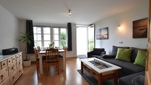 A modern living room with a dining area, a sofa, a coffee table, and a large window. - App.7 / 3-room (5 adults + 1 infant), ground floor - Ferienwohnungen Zum Breitling OVS 691 (Insel Poel)