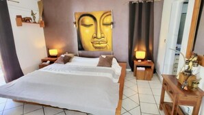 A bedroom with a large bed, two bedside tables, a wall-mounted Buddha statue, and a wooden nightstand with a lamp. - Studio Boudha Zen   Residence Adam et Eve (Baie Orientale)