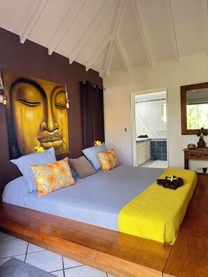 Une chambre avec un grand lit, un plaid jaune et un tableau représentant un Bouddha accroché au mur.