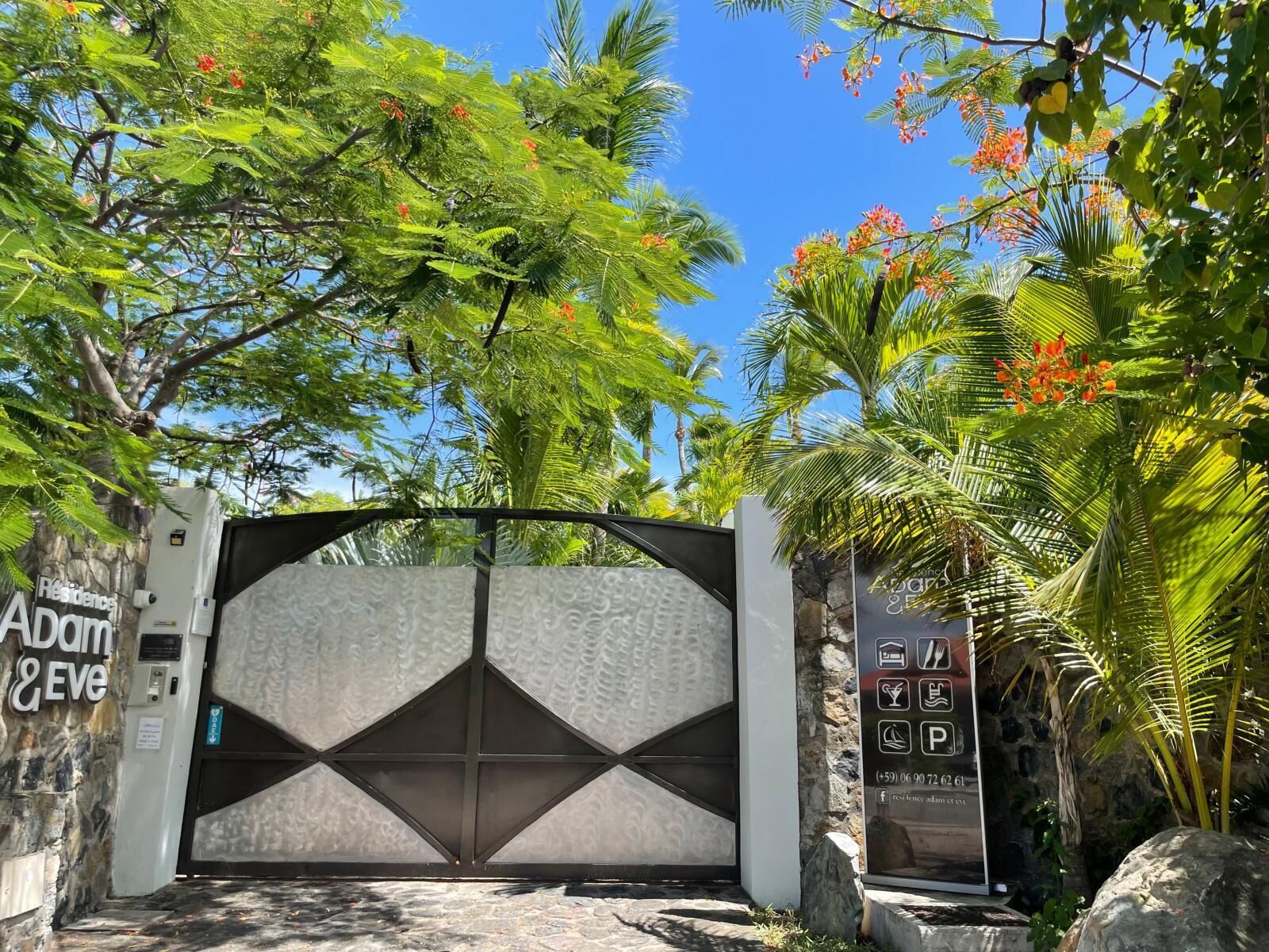 A gated entrance with a geometric design, a stone wall, and a signboard with amenities and contact information.