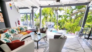A modern outdoor living area with a white sofa, a glass table, and a white armchair. There is a view of a pool and palm trees in the background. - Studio Boudha Zen   Residence Adam et Eve (Baie Orientale)