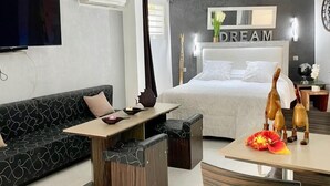 A modern hotel room with a sofa, a dining table, a bed with a 'DREAM' sign, and a wall-mounted TV.