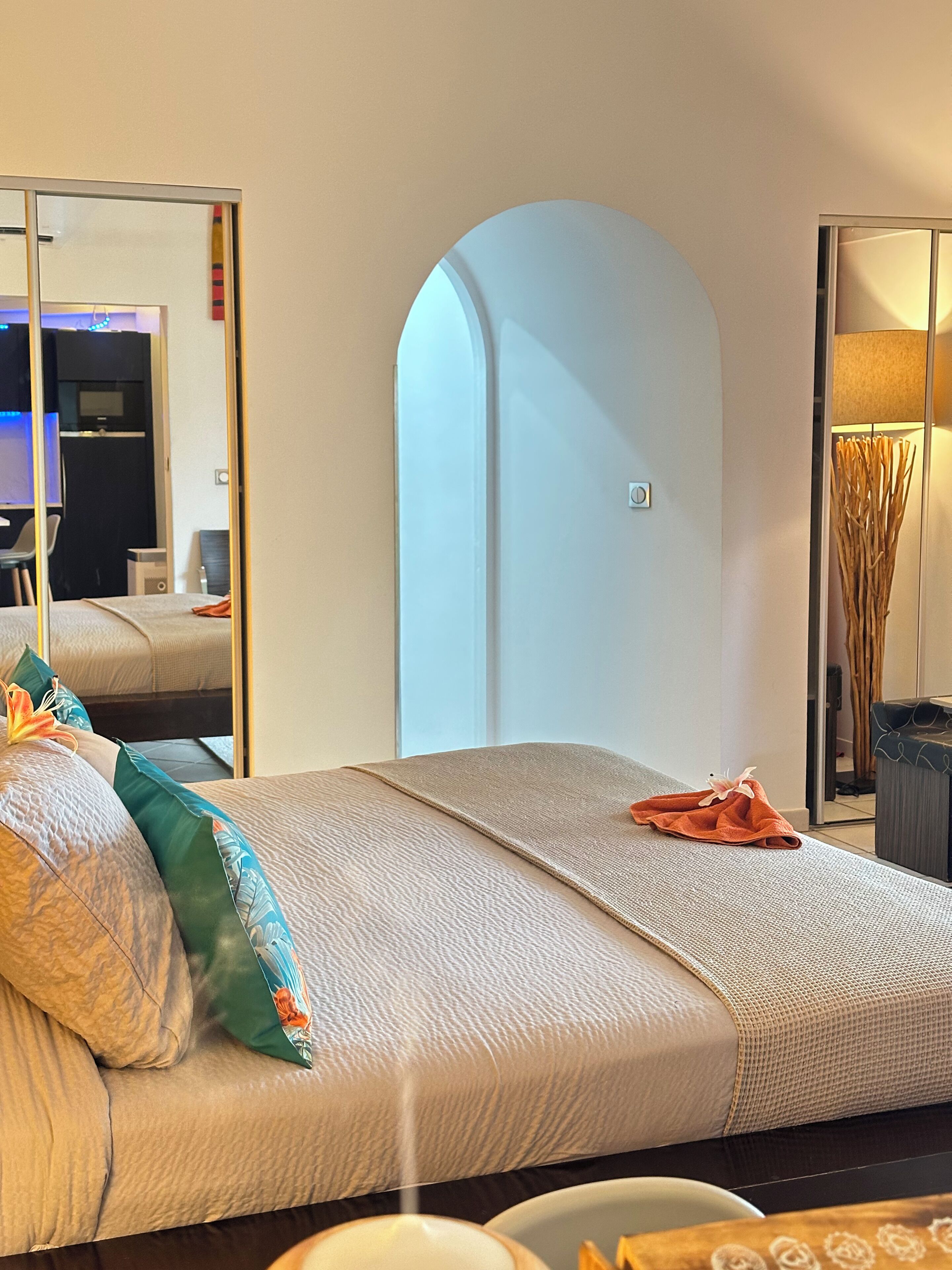 A bed with a headboard, pillows, and a blanket. There is a mirror on the wall and a lamp beside the bed.