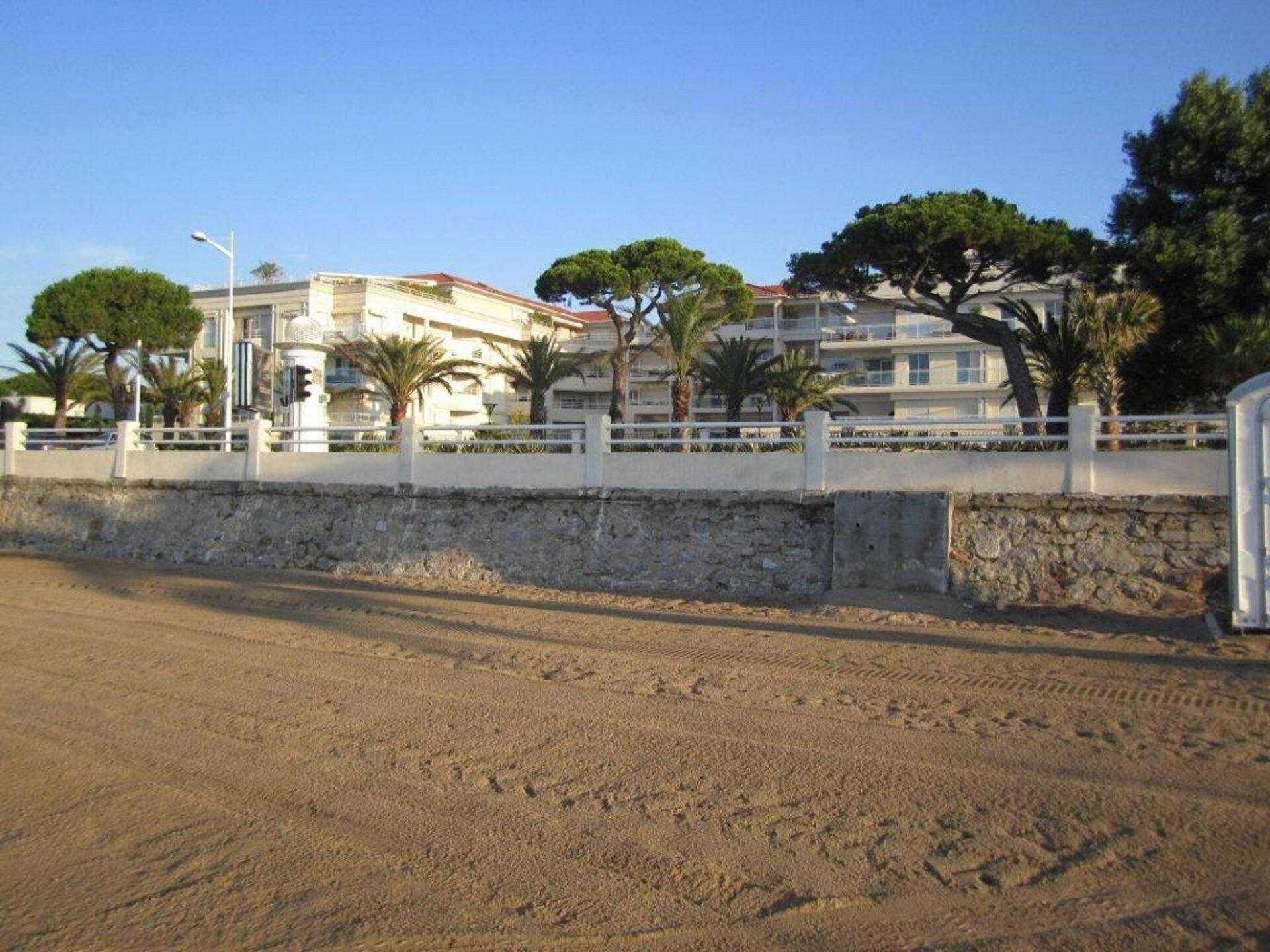 Une route côtière bordée d’un mur de pierres et d’une clôture blanche. On aperçoit des bâtiments dotés de balcons et des arbres en arrière-plan.