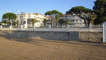 Une route côtière bordée d’un mur de pierres et d’une clôture blanche. On aperçoit des bâtiments dotés de balcons et des arbres en arrière-plan.