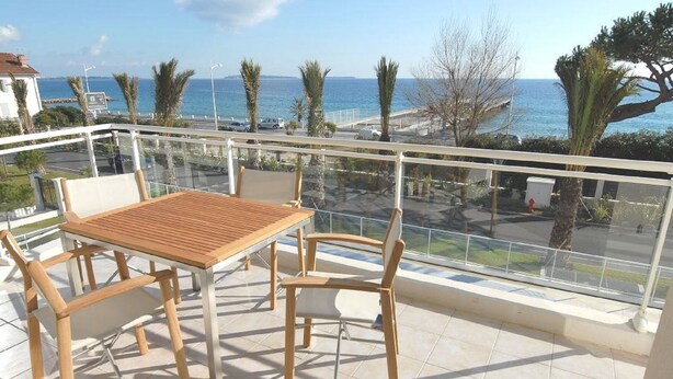 A balcony with a wooden table and chairs, overlooking a coastal area with palm trees and a clear view of the sea. - Royal Palm 218, with panoramic sea view (Cannes)