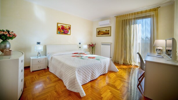 A bedroom with a wooden floor, a bed with a floral quilt, a bedside table with a lamp, a desk with a chair, and a window with curtains.