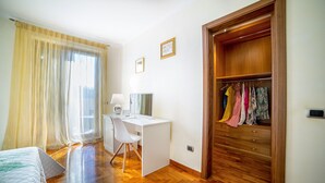 A bedroom with a wooden floor, a desk with a chair, a bed, and a wardrobe with hanging clothes.