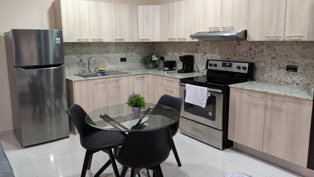 A modern kitchen with light wood cabinets, a stainless steel refrigerator, a gas stove, and a glass dining table with chairs. - Hotel Suites Merlyn Airport (San Juan)