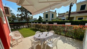 Une terrasse extérieure avec une table ronde et des chaises, entourée d’un jardin agrémenté de haies et de fleurs.