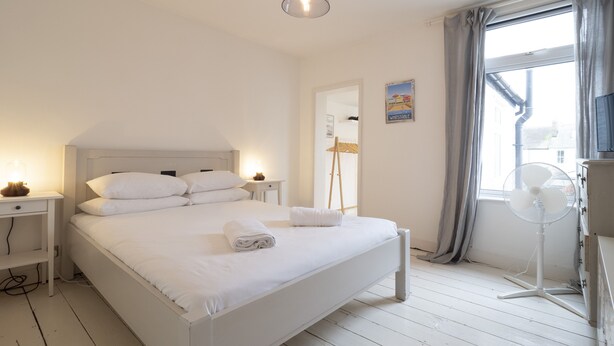 A white bedroom with a bed, bedside tables, a standing fan, a window with curtains, and a television. - Crab Cottage (Whitstable)