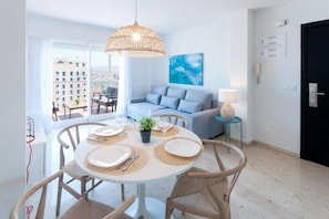 A modern living room with a round dining table set for four, a sofa, and a large window offering a city view. - AG Calmo Singular Apartments (Gandia)