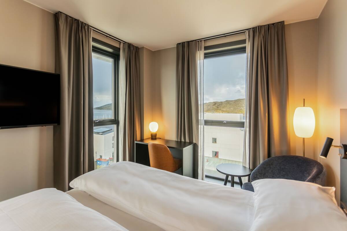 A hotel room with a bed, a desk with a chair, a TV mounted on the wall, and large windows with curtains.