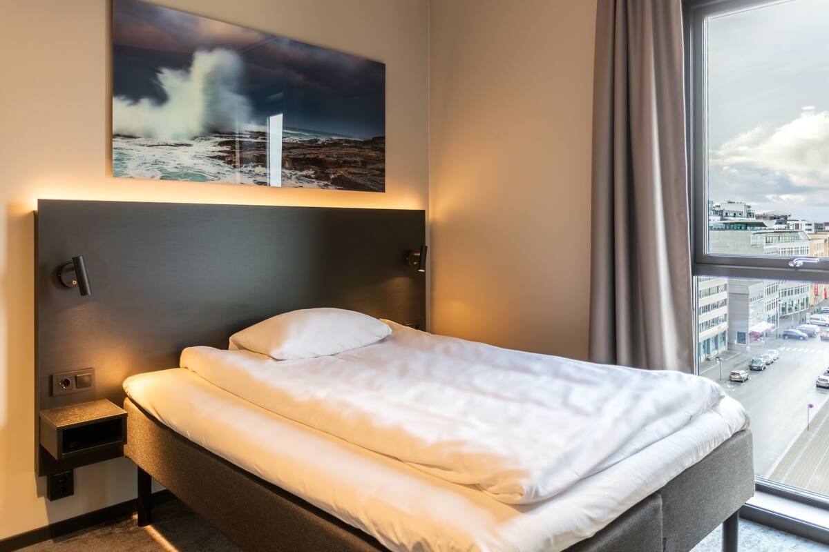 A hotel room with a bed, a headboard, a nightstand, a window with a city view, and a painting of a stormy sea.