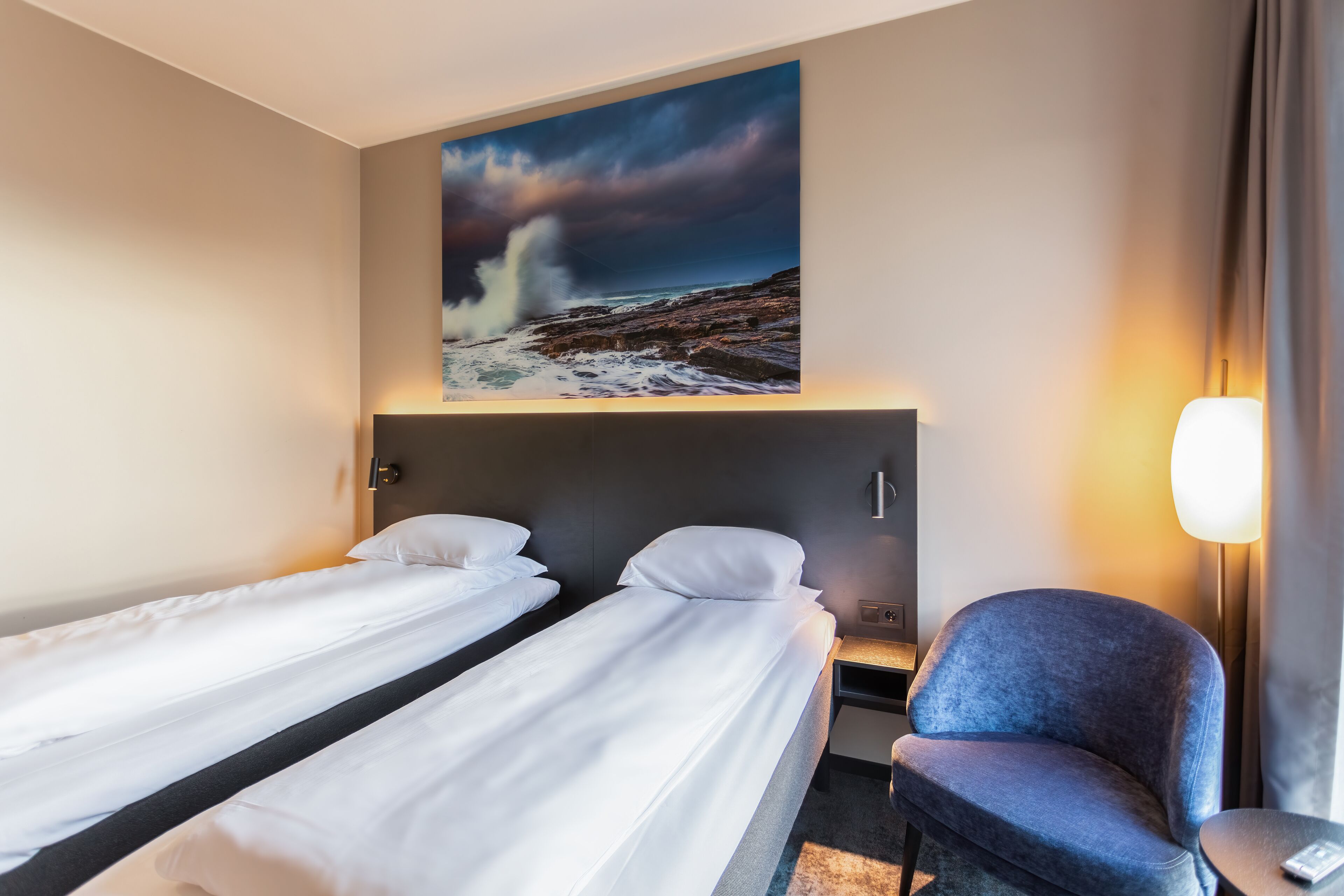 Habitación de hotel con dos camas, una silla, un cuadro de un mar embravecido y una lámpara.