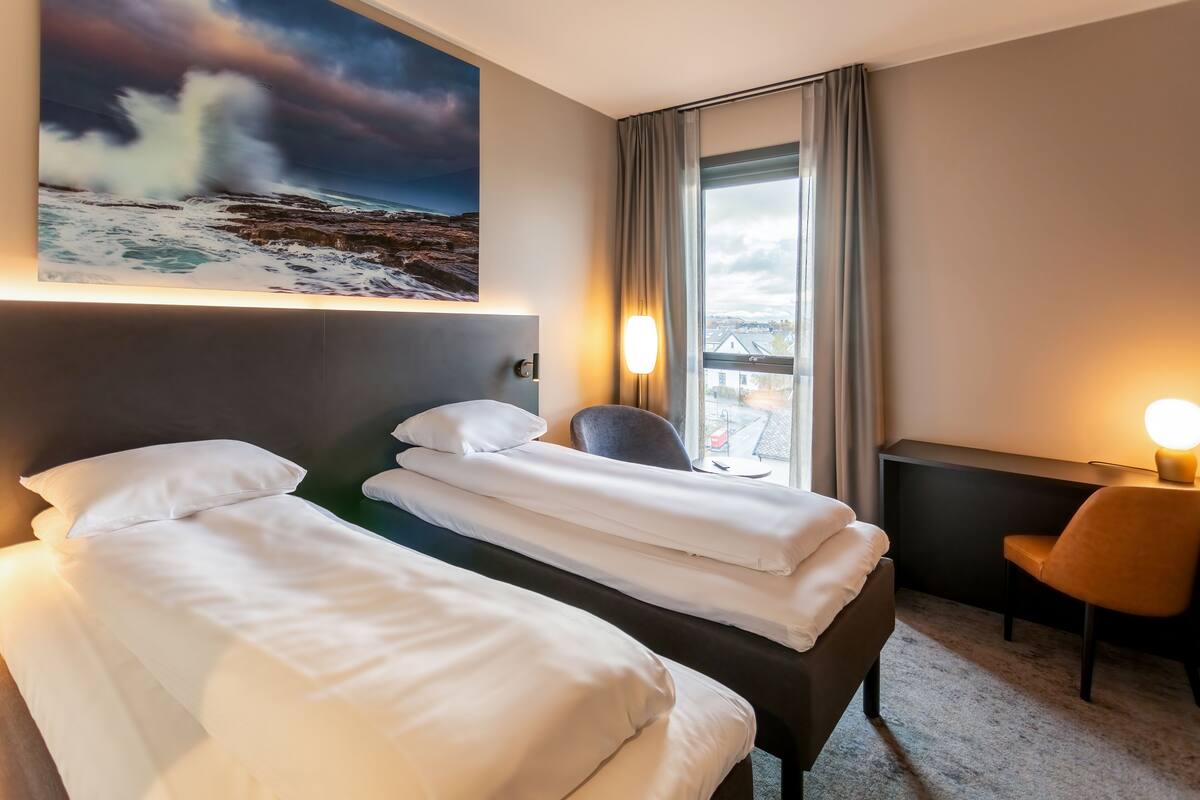 A hotel room with two beds, a desk, a chair, a large window with a view, and a painting of a seascape.
