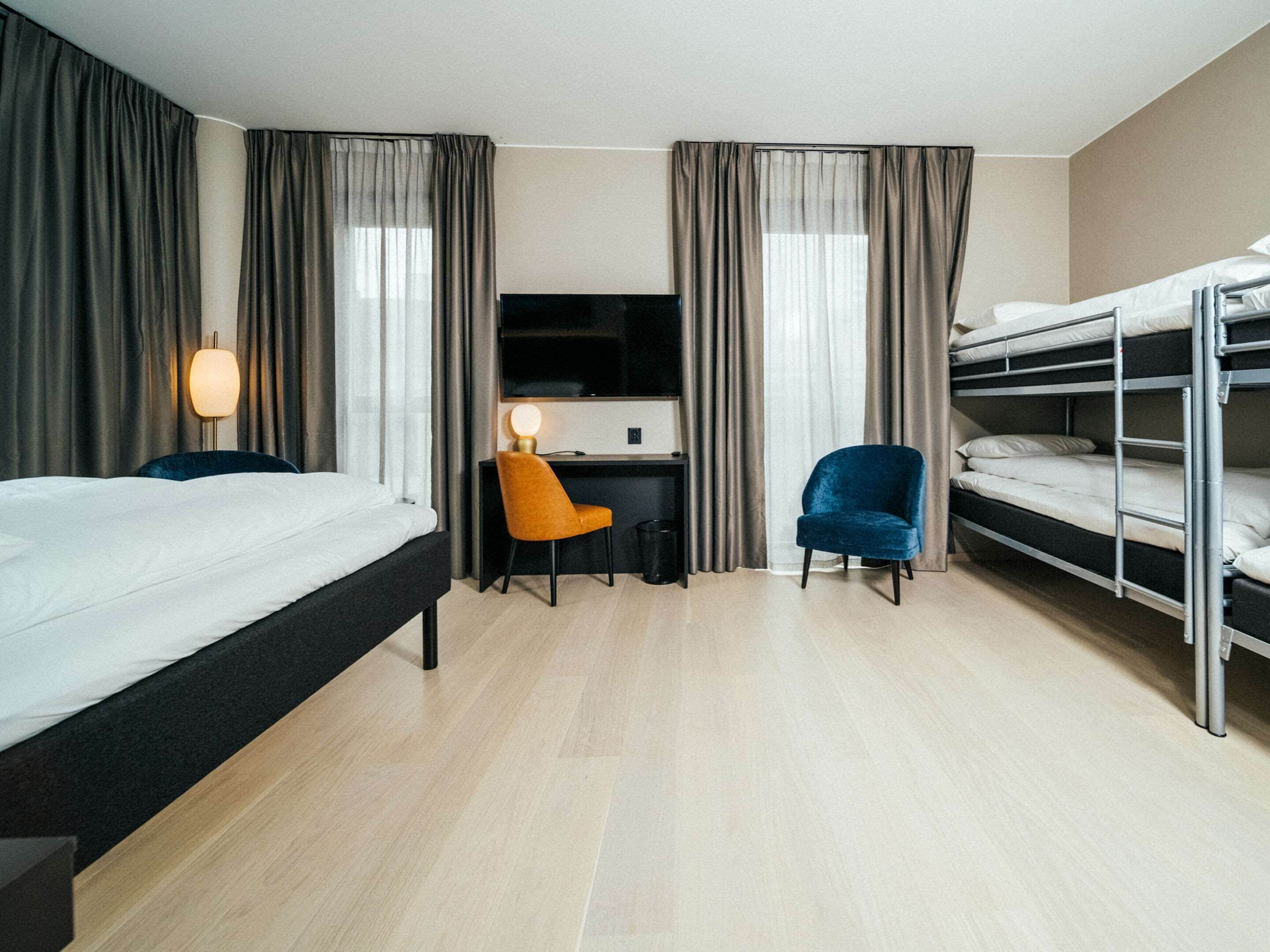 A hotel room with a bed, desk, chair, bunk beds, and curtains.