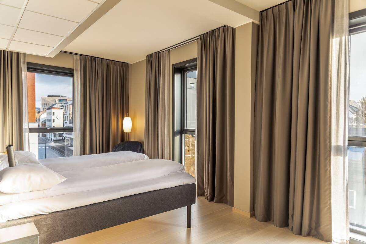 A modern hotel room with a large bed, a nightstand, a lamp, and a view of the city through large windows.
