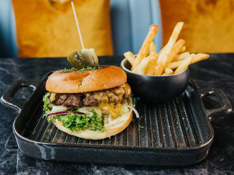 A grilled burger with a bun, lettuce, and pickles on a black skillet, accompanied by a small bowl of fries.