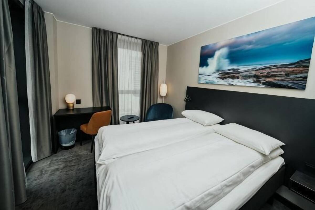 A hotel room with a bed, a desk with a chair, a small table, and a painting on the wall.
