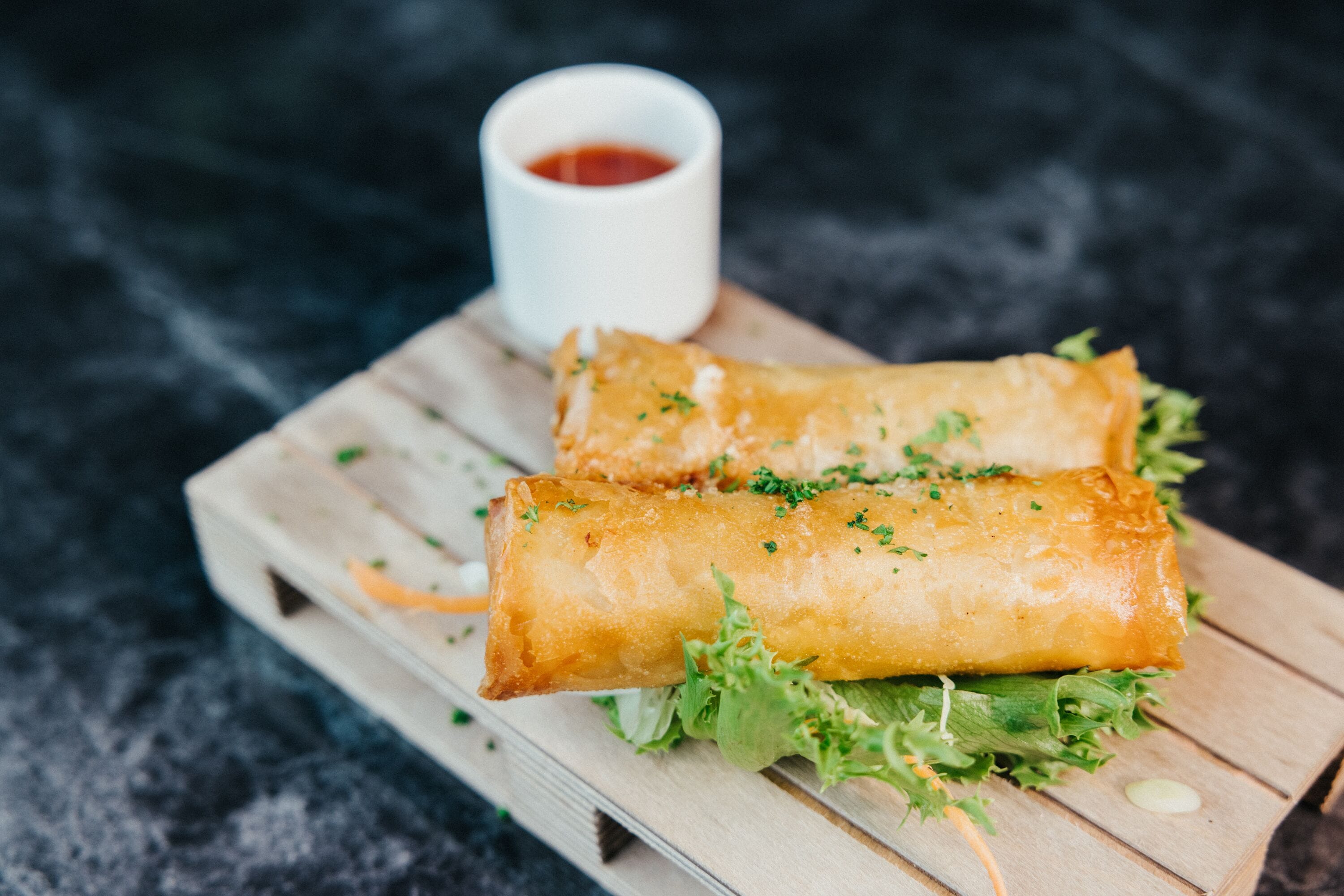 Two spring rolls on a wooden board with lettuce and a small cup of sauce.
