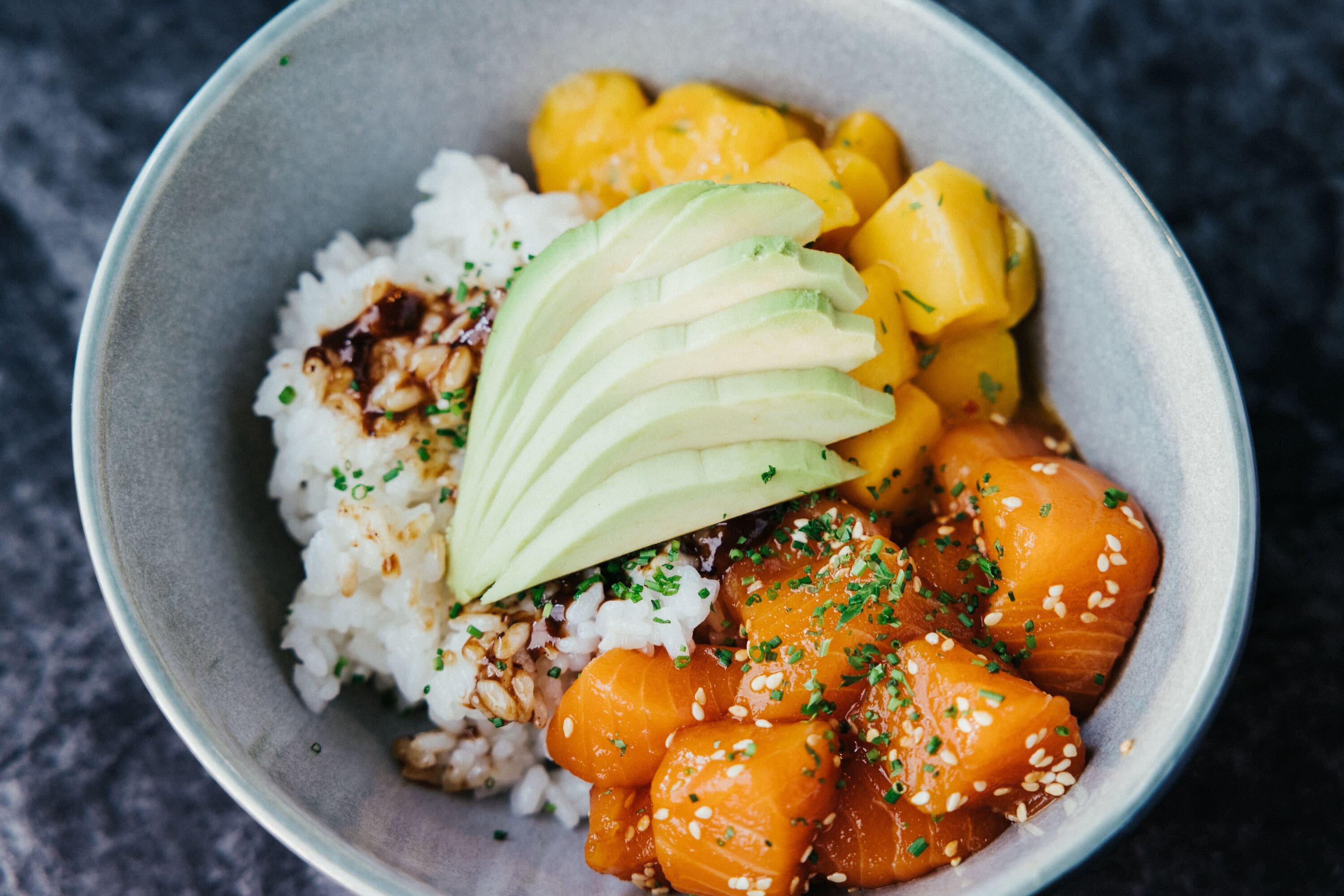 A bowl of food with rice, avocado slices, mango chunks, and salmon, garnished with sesame seeds and chopped herbs.