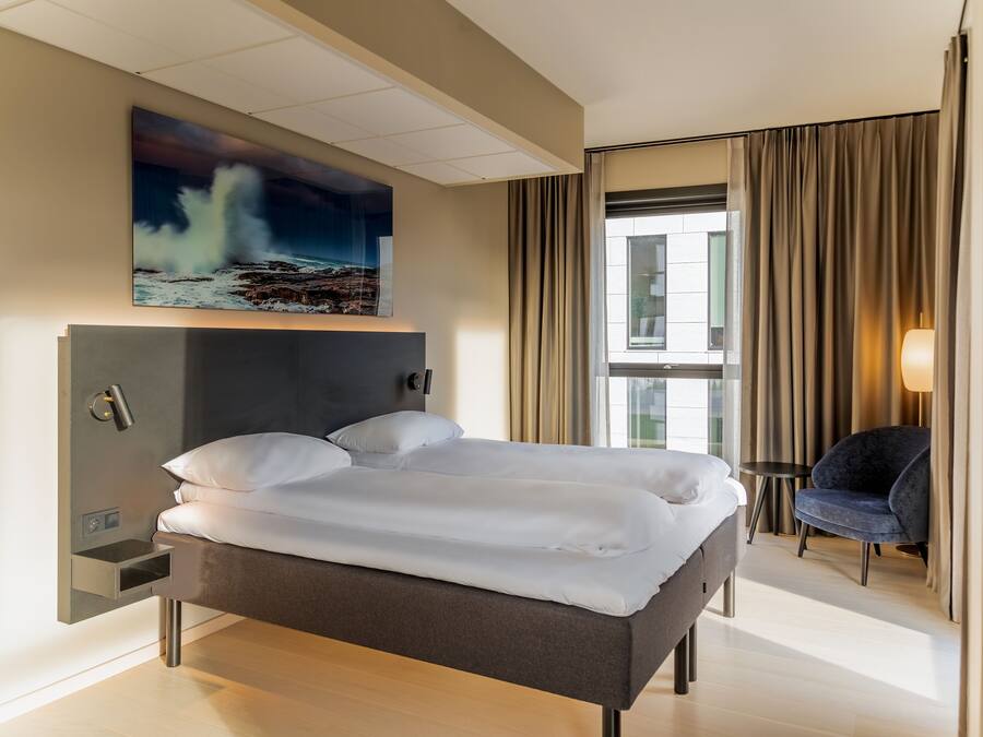 Una habitación de hotel moderna con una cama, una silla, un cuadro y un ventanal con cortinas.