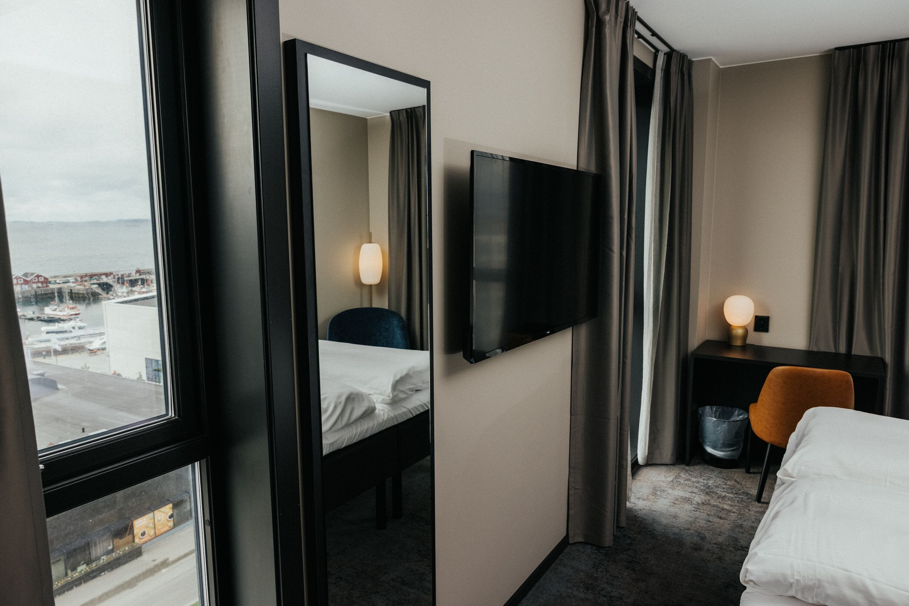 A hotel room with a bed, a desk with a chair, a TV mounted on the wall, and a window offering a view of the city.