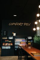 Un interior oscuro con un letrero de neón que dice "COMFORT DELI", estantes llenos de aperitivos y bebidas, y una zona de bar.