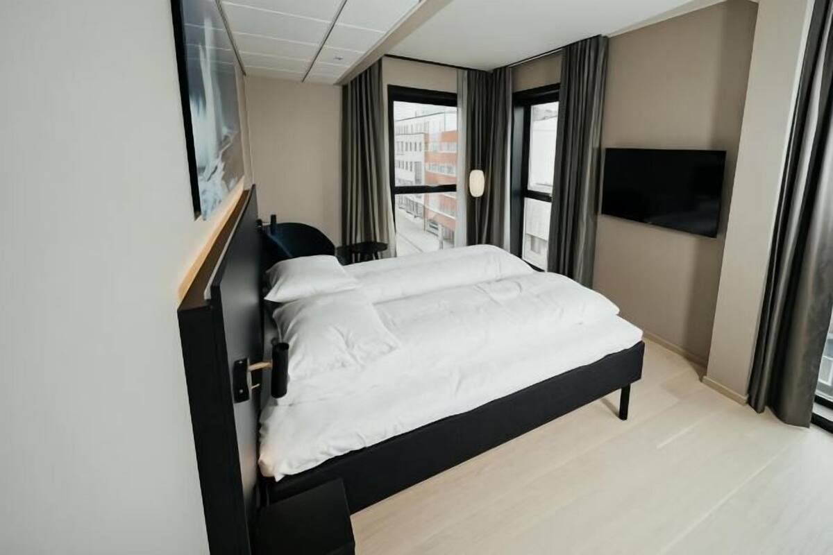 A modern hotel room with a bed, a TV mounted on the wall, and a window with curtains.