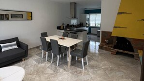 Un espacio habitable moderno con comedor, un sofá negro, una mesa blanca y una cocina al fondo.