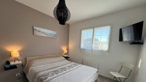 Un dormitorio con cama, dos lámparas de noche, un radiador, una ventana con cortinas, un televisor montado en la pared y una silla plegable.