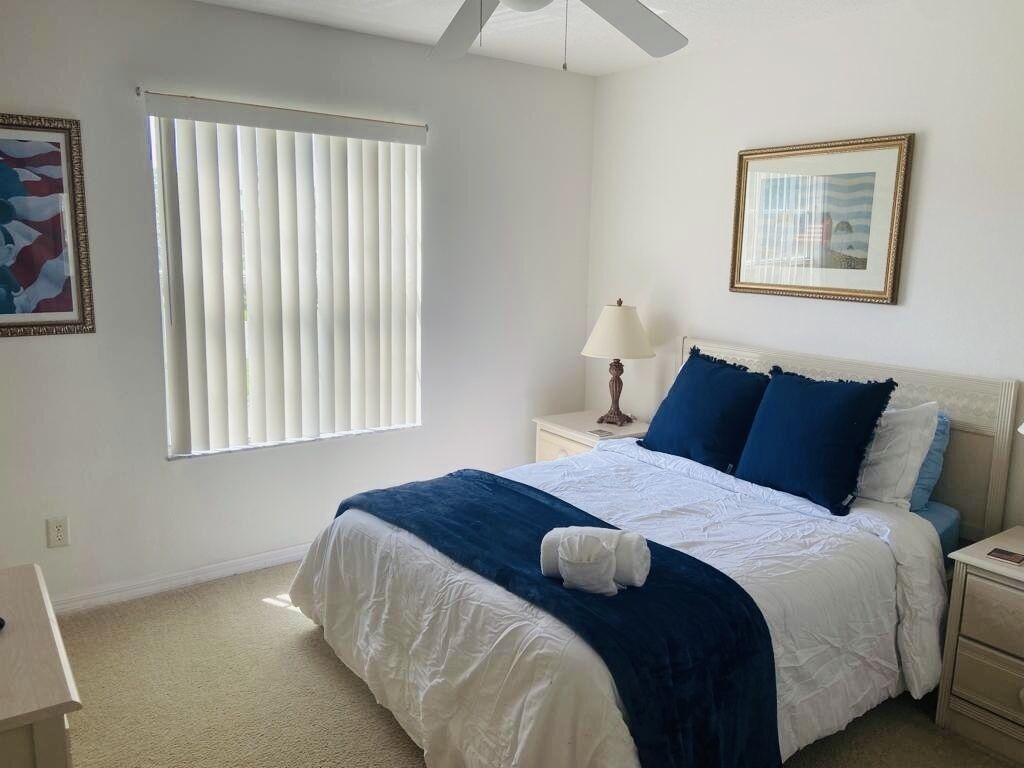 Une chambre à coucher comprenant un lit, une table de chevet, une lampe, une fenêtre avec des stores et un tableau encadré accroché au mur.