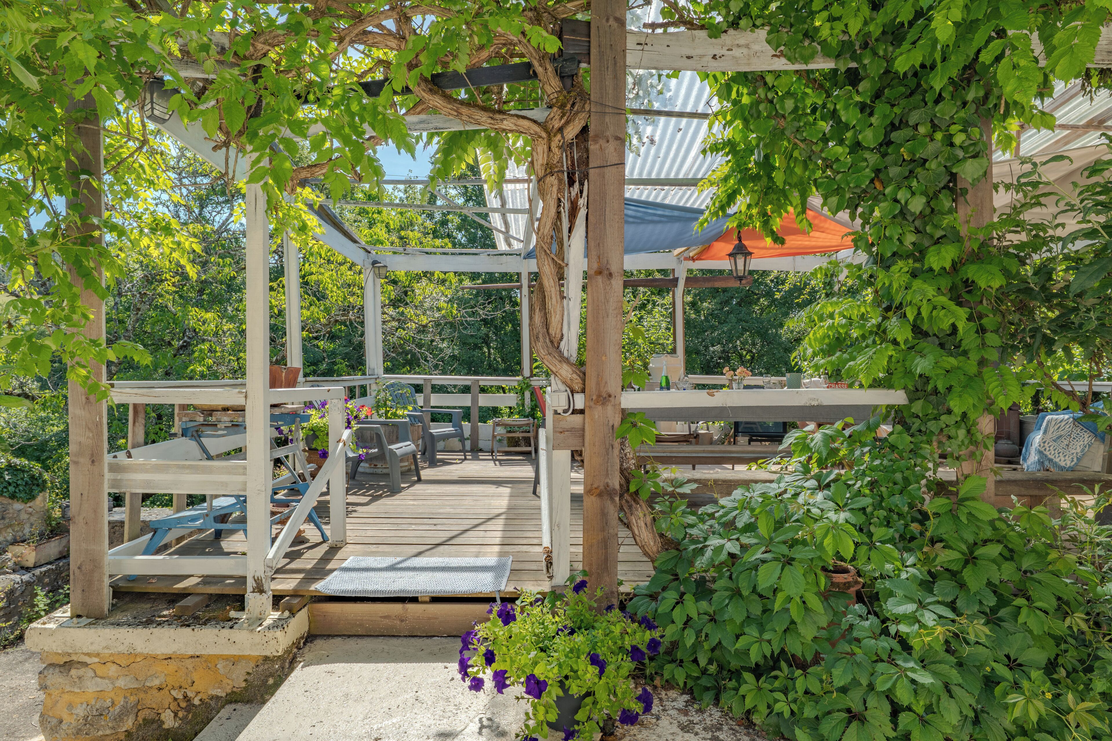 Um deck de madeira com assentos, cercado por vegetação e uma estrutura de pérgola.